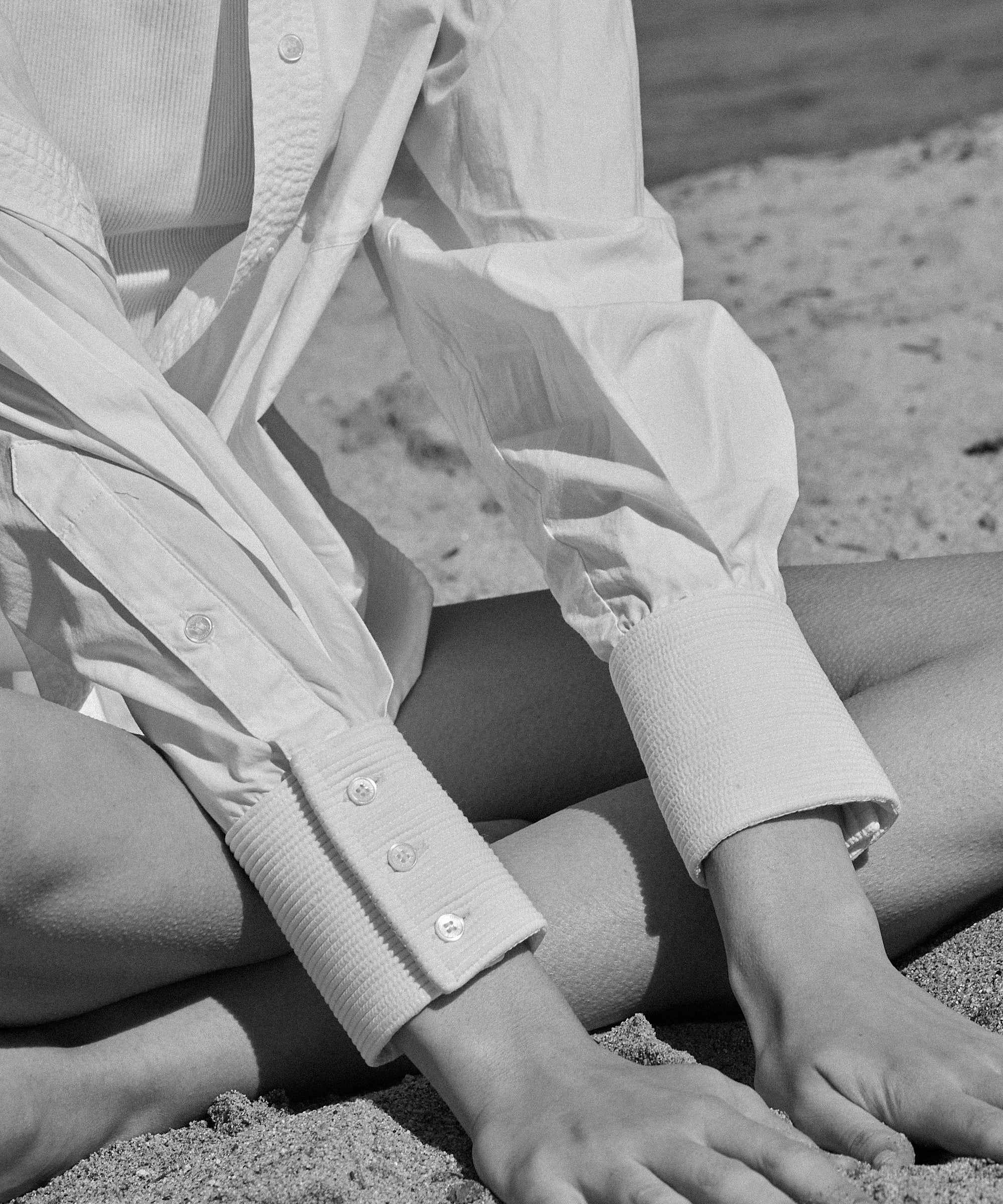 Woman in white cotton poplin ELE shirt with hand-embroidered details sitting on sand, luxury minimalist fashion