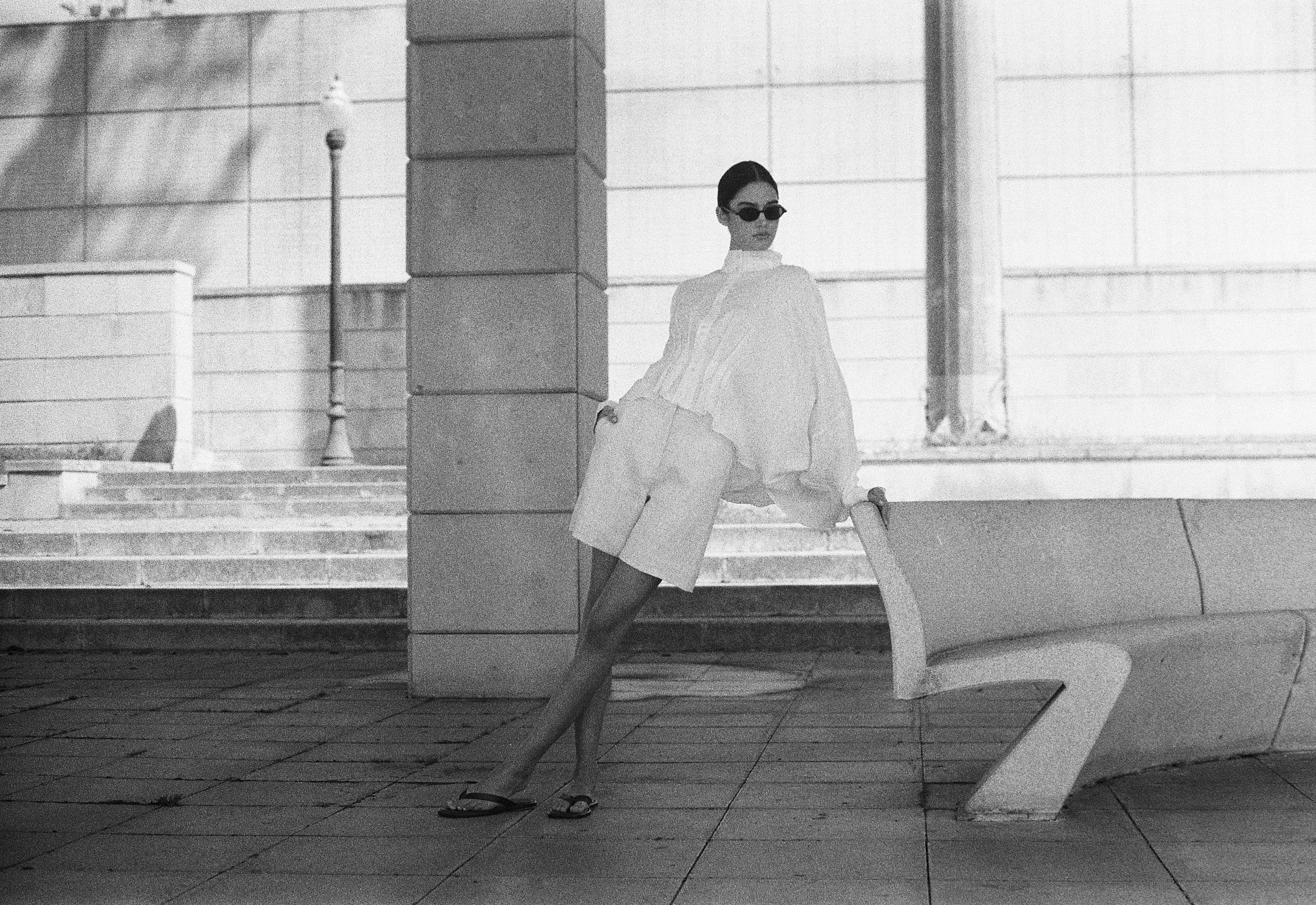 Woman in cream linen shirt and tailored trousers with round sunglasses, seated on architectural concrete bench in minimalist urban setting