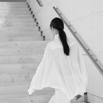 Woman wearing white Italian linen CATE shirt climbing stairs in minimalist stairwell, embodying Aymone Studio's timeless luxury essentials