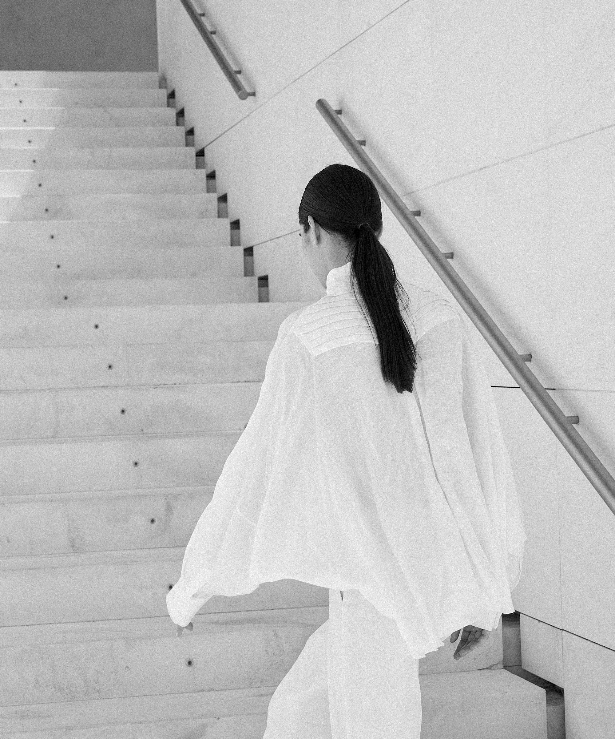 Woman wearing white Italian linen CATE shirt climbing stairs in minimalist stairwell, embodying Aymone Studio's timeless luxury essentials