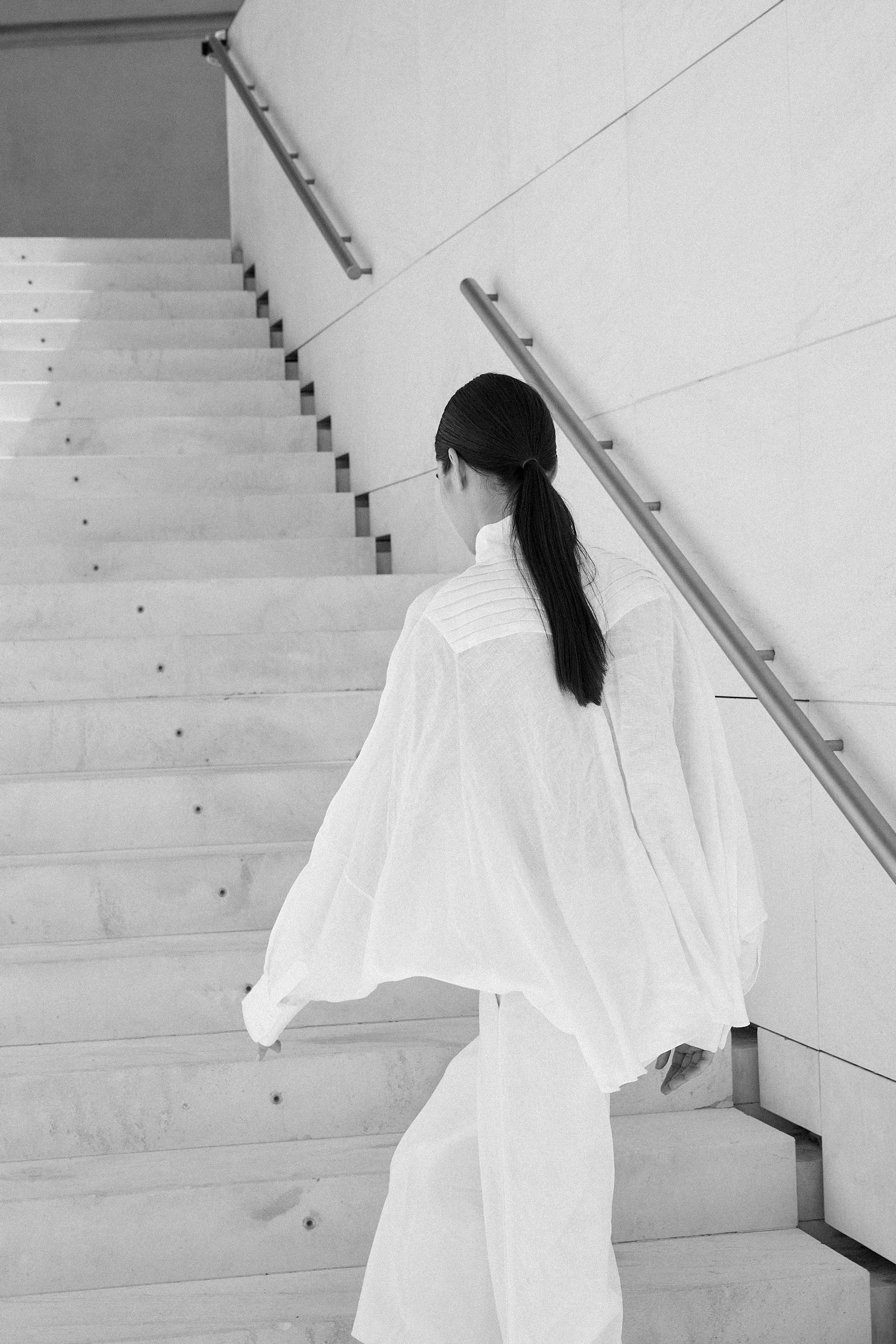 Woman wearing white Italian linen CATE shirt climbing stairs in minimalist stairwell, embodying Aymone Studio's timeless luxury essentials